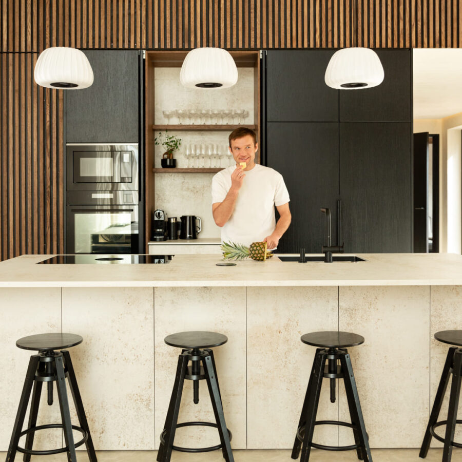 Smiling man standing at a modern kitchen island with wooden accents, eating a slice of pineapple.