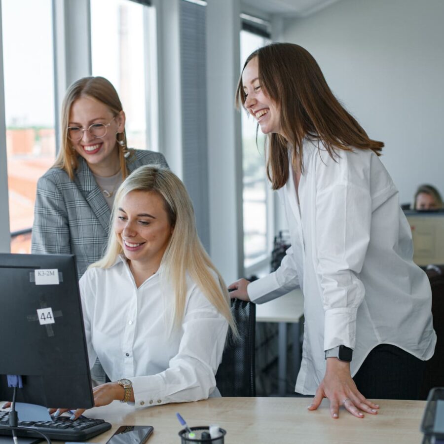 A business team collaborating in a contemporary office setting, featuring three women smiling and actively engaged around a computer, fostering a positive and dynamic work atmosphere.