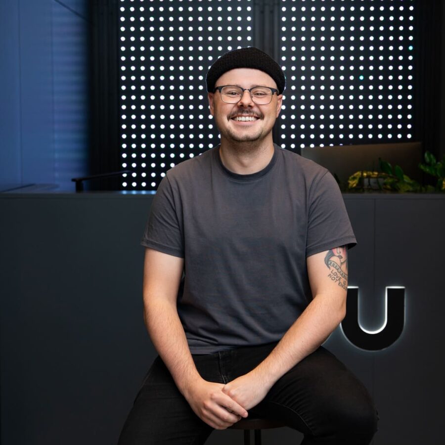 Friendly team member in casual attire and beanie, seated in a modern office lobby with sleek LED panel and greenery backdrop, showcasing a welcoming workplace culture.